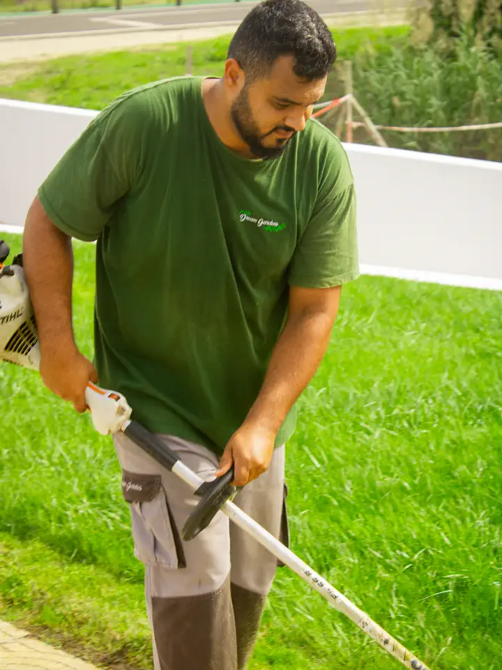 Jardineiro a cortar relvado com uma roçadora, junto a um muro branco