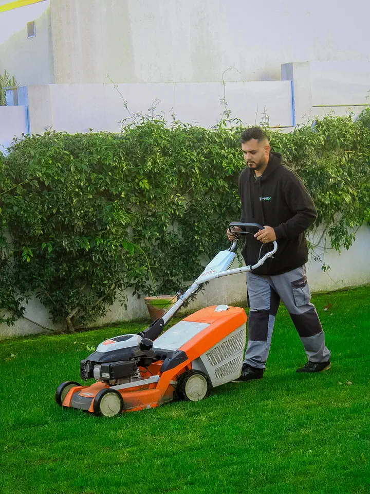 Jardineiro a cortar relvado com um corta-relvas laranja, junto a uma sebe e a um muro branco